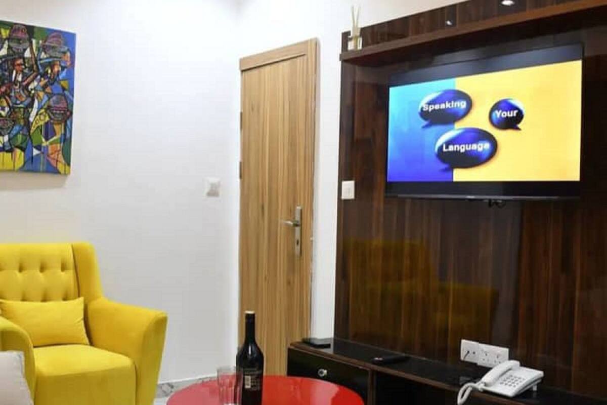 Una habitación moderna con un sillón amarillo, una mesa roja, una botella de vino, una pantalla de televisión mostrando contenido para aprender idiomas y un armario de madera.