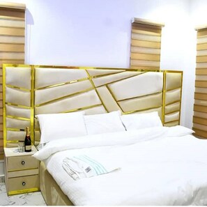 A bed with a gold headboard, white bedding, and a bottle of champagne on a bedside table. - Tito Empire (Lekki)