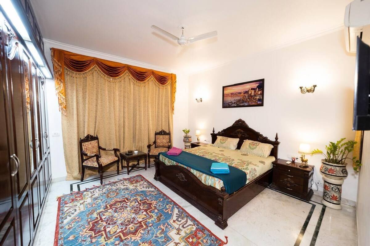 A bedroom with a large bed, a desk, two chairs, a ceiling fan, a nightstand, a potted plant, and a wall-mounted TV.