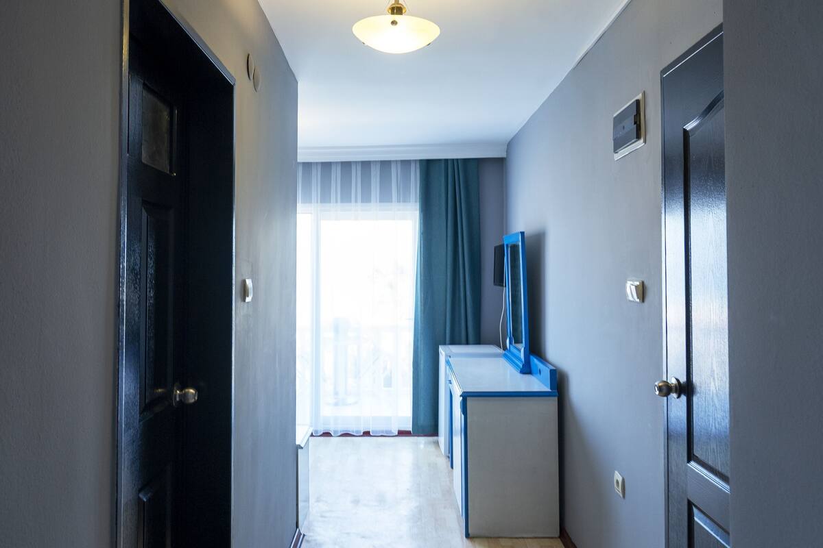 Um corredor com piso de madeira, uma porta à esquerda, uma pequena escrivaninha com um objeto azul sobre ela e uma janela com cortinas ao fundo.