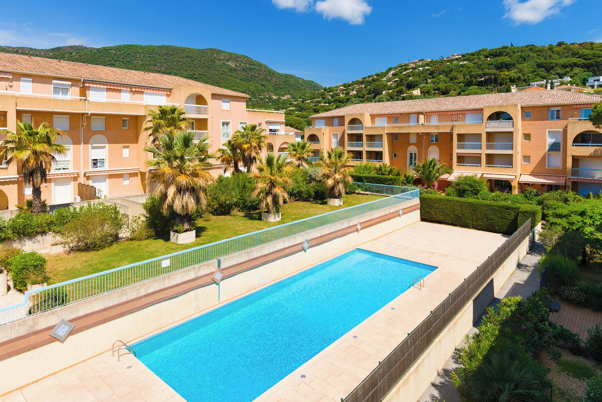 A swimming pool surrounded by a building with balconies, palm trees, and a hilly landscape in the background.