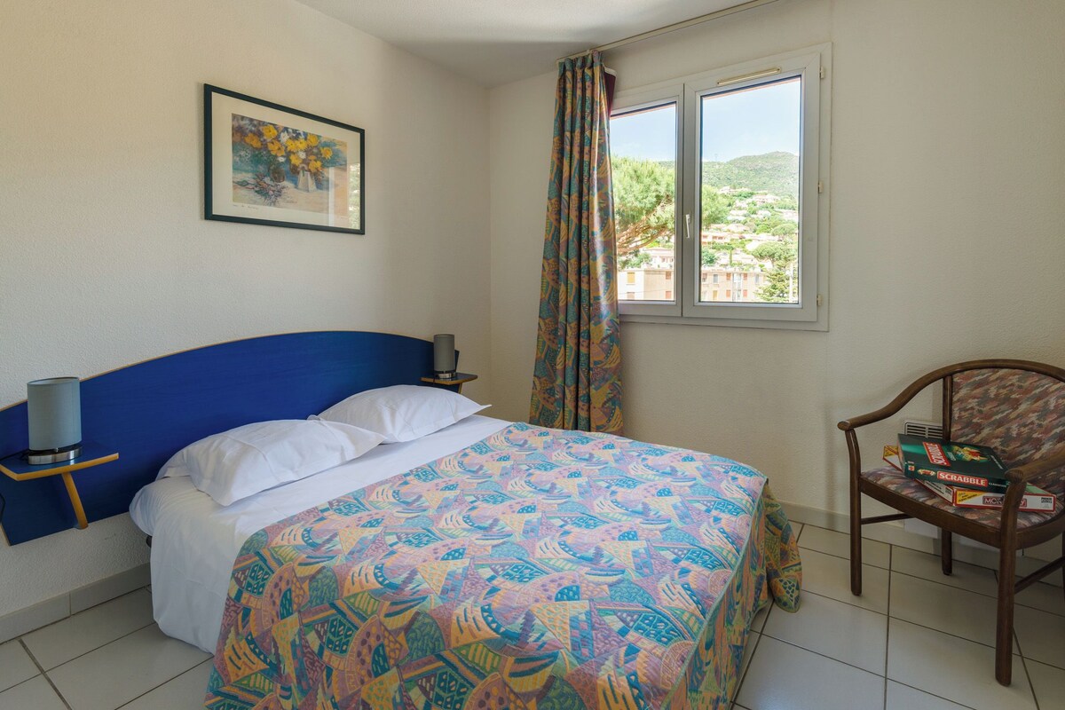 Un dormitorio con cama, una silla, un ventanal con cortinas, una mesita de noche y un cuadro enmarcado en la pared.