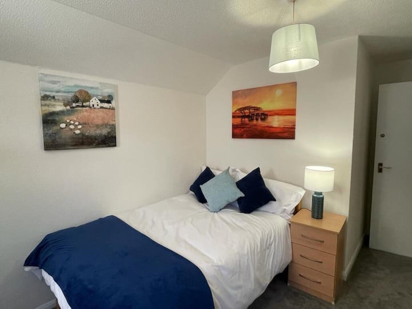 A bedroom with a bed, a bedside table with a lamp, and two wall-mounted paintings.
