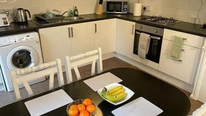 A compact kitchen with white cabinets, a microwave, a stove, and a washing machine. There is a dining table with a bowl of fruit and napkins.