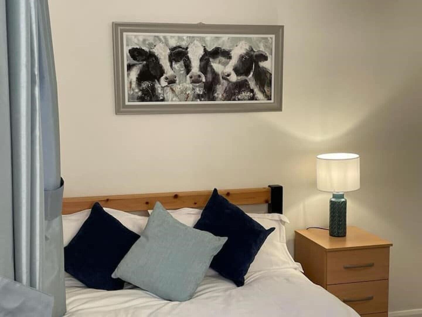 Quarto com cama, almofadas, mesa de cabeceira com abajur e um quadro na parede.