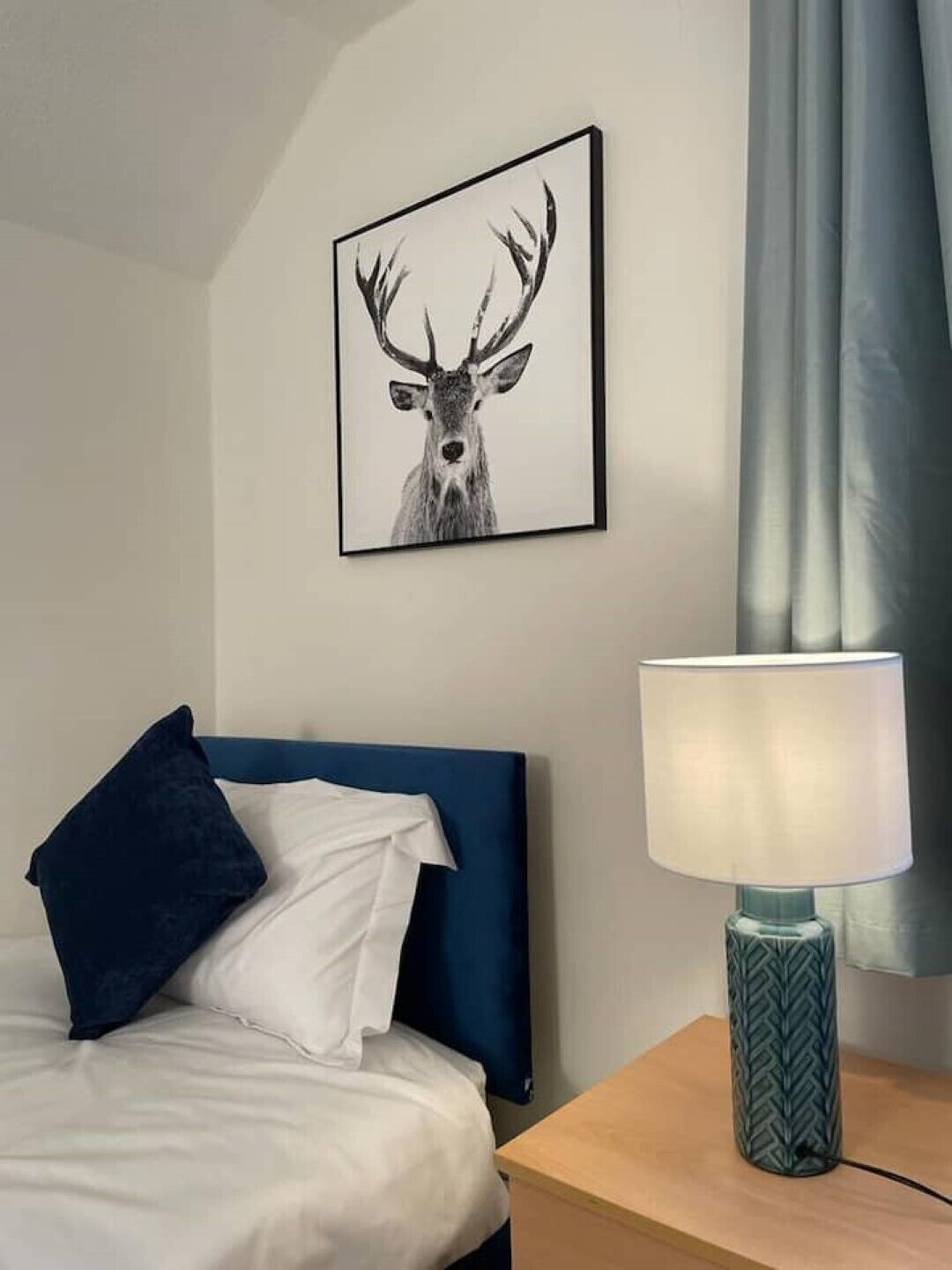 Een hotelkamer met een bed, een blauw hoofdeinde, een wit kussen, een donkerblauw kussen, een afbeelding van een hert in een lijst aan de muur en een lamp op een houten nachtkastje.