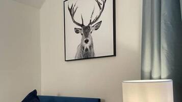 Een hotelkamer met een bed, een blauw hoofdeinde, een wit kussen, een donkerblauw kussen, een afbeelding van een hert in een lijst aan de muur en een lamp op een houten nachtkastje.