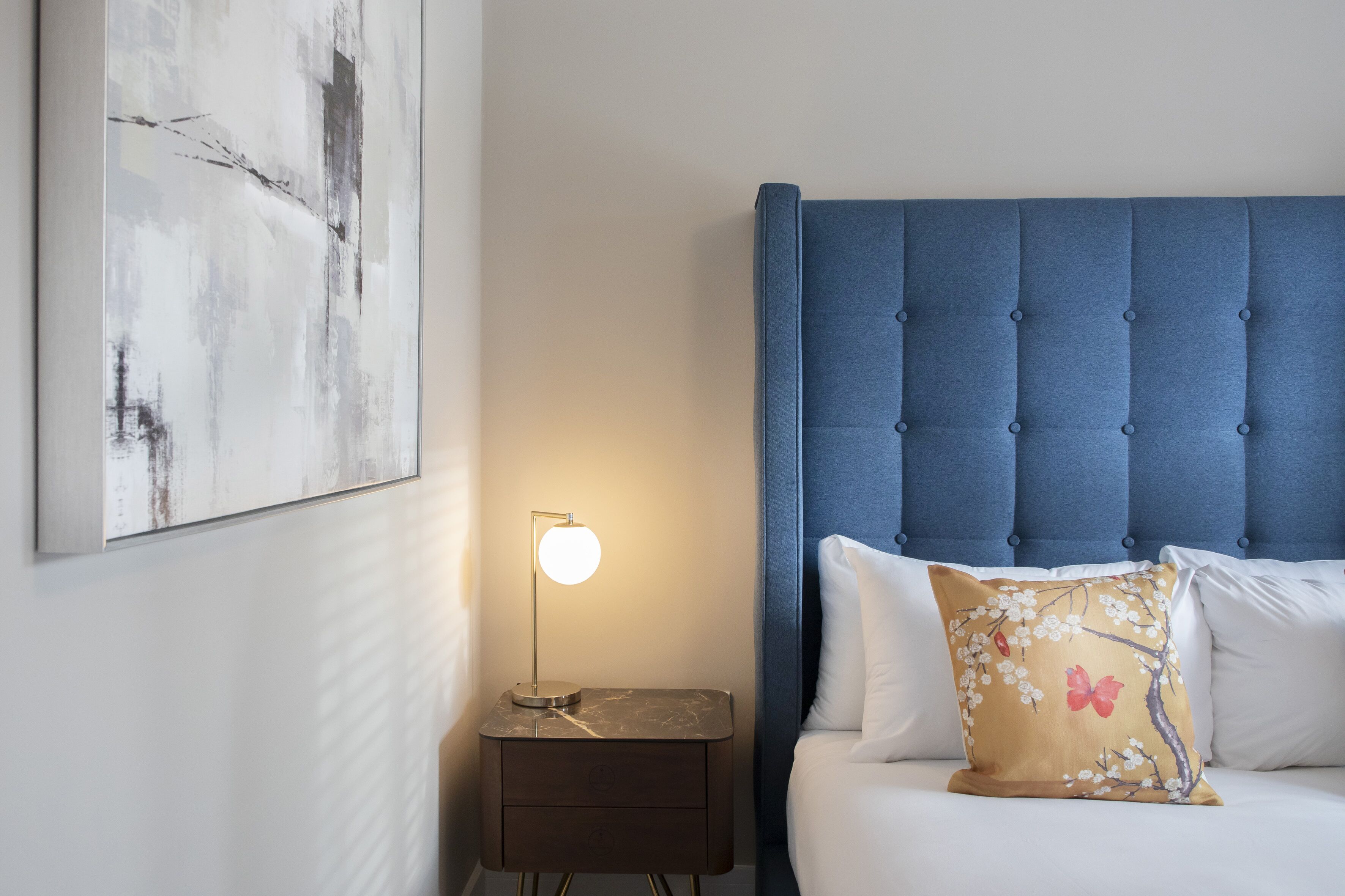 Een slaapkamer met een gestoffeerd hoofdeinde, een nachtkastje met een lamp en een bloemenkussen.