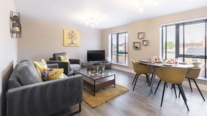 A modern living room with a sofa, coffee table, and dining area with chairs. A flat-screen TV is mounted on the wall, and there are windows with blinds. - Elliot Oliver - Luxurious 2 Bedroom Apartment With Parking (Gloucester)