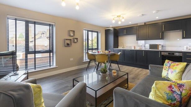 A modern kitchen with dark cabinets and a dining area with a glass table. - Elliot Oliver - Luxurious 2 Bedroom Apartment With Parking (Gloucester)