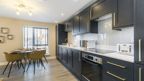 A modern kitchen with dark cabinetry, a white countertop, and a dining area with yellow chairs. - Elliot Oliver - Luxurious 2 Bedroom Apartment With Parking (Gloucester)