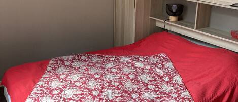 A bedroom with a bed, a wardrobe, and a small shelf with a fan.