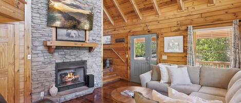 A cozy cabin interior with a stone fireplace, wooden beams, a sofa, and a ceiling fan.