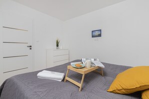 A neatly arranged bedroom with a bed, a bedside table with a tray holding a book, a cup, and a pastry, a bedside plant, and a painting on the wall.