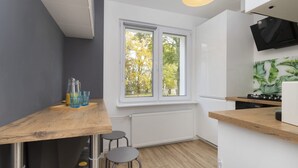A modern kitchen with a wooden table, two stools, a window with a view of trees, a white radiator, and a black TV mounted on the wall.