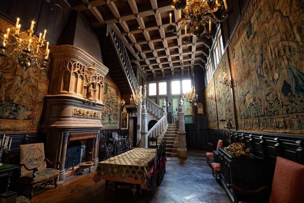 A grand hall with a high ceiling, ornate chandeliers, a fireplace, and a staircase leading to an upper level.
