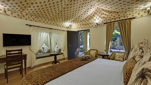 A hotel room with a large bed, a television mounted on the wall, a window with curtains, a small table with a chair, and a door leading outside. -  GIR LION SAFARI CAMP (Mendarda)