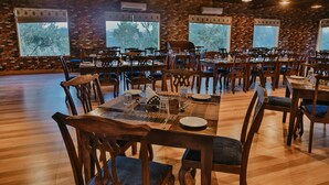 A spacious dining area with multiple tables and chairs, brick walls, and large windows offering views of trees. -  GIR LION SAFARI CAMP (Mendarda)