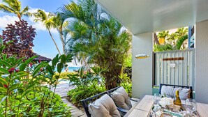 A patio with a table set for two, surrounded by lush tropical plants and a view of the ocean. - Upgraded beachside condo with ground floor access (Lahaina)