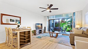 A spacious living area with a wooden dining table, chairs, a sofa, and a TV. There is a ceiling fan, a large window with a view of greenery, and a sliding glass door leading to an outdoor area. - Upgraded beachside condo with ground floor access (Lahaina)