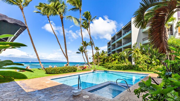 A pool area with a view of the ocean, surrounded by palm trees and a building with balconies. - Upgraded beachside condo with ground floor access (Lahaina)