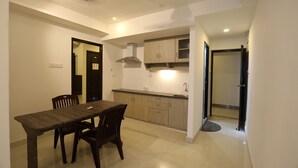 A dining area with a wooden table and chairs, a kitchen with wooden cabinets and a sink, and a door leading to another room. - Hotel Rydham (Madurai)