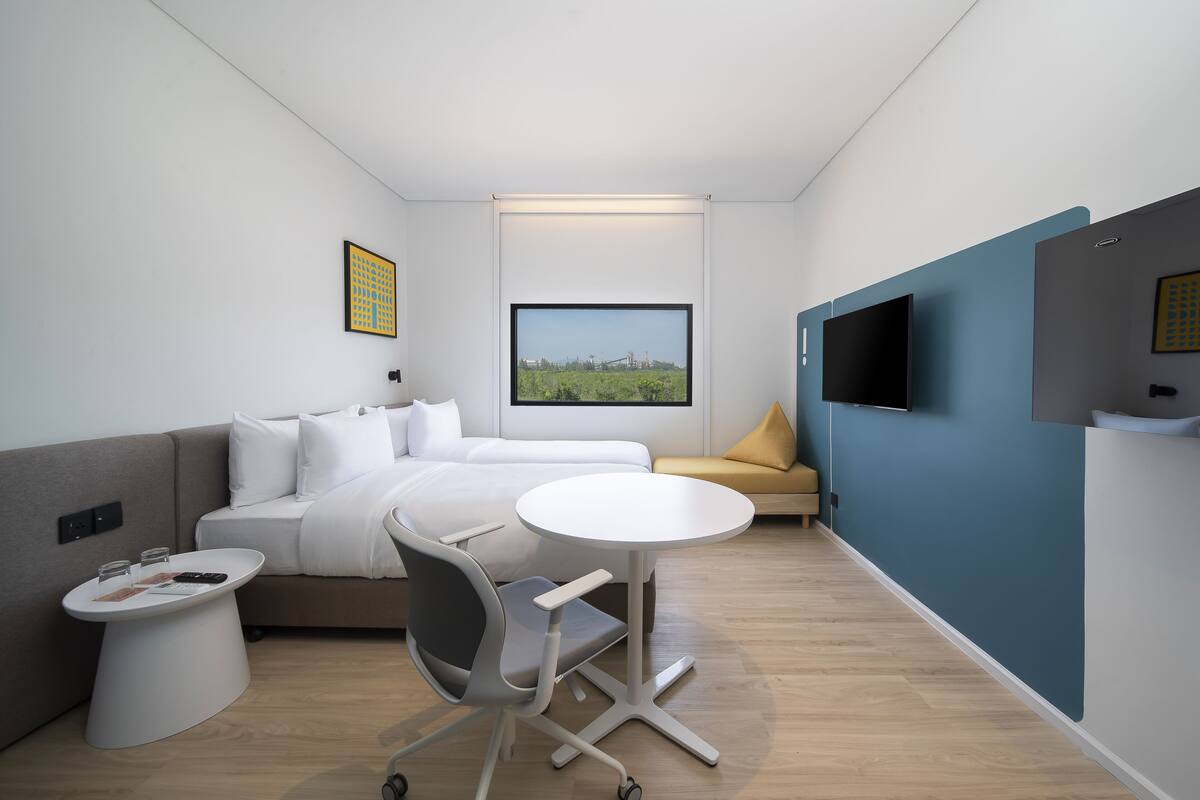 A modern hotel room with a bed, a sofa, a small round table, a chair, a TV, and a large window with a city view.