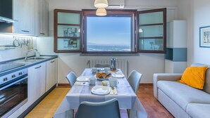 Un comedor con una mesa dispuesta para cuatro, una cocina con electrodomésticos modernos, un ventanal con vistas y una zona de estar acogedora con un sofá y un cojín amarillo.