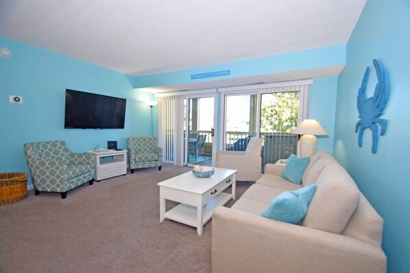 A living room with a TV, two patterned chairs, a white coffee table, and a beige sofa with blue cushions.