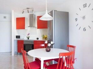 Una cocina moderna con armarios rojos, una mesa de comedor blanca con sillas rojas, una lámpara colgante blanca y un reloj de pared.