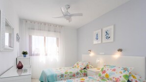 Une chambre avec deux lits, un ventilateur de plafond, une table de chevet avec une lampe et une fenêtre avec des rideaux légers.
