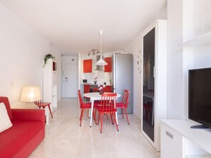 Una sala moderna con un sofá rojo, una mesa de comedor con sillas rojas, un televisor sobre un mueble blanco y una cocina al fondo.