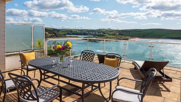 Eine Terrasse mit Tisch und Stühlen mit Blick auf eine Küstenlandschaft mit Hügeln und Strand.