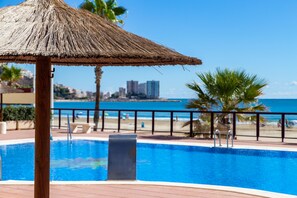 Un área de piscina con una sombrilla de paja, vistas a la playa y un paisaje urbano al fondo.