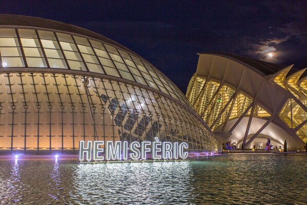 A large, modern building with a distinctive, curved glass facade and a reflective water surface in front.