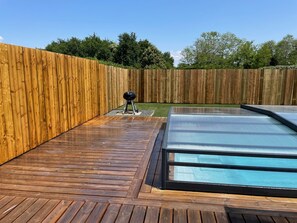 Una terraza de madera con piscina cubierta de vidrio y una parrilla.