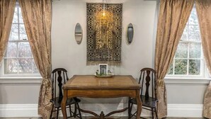 A dining room with a wooden table and four chairs, a chandelier, and two windows with curtains.