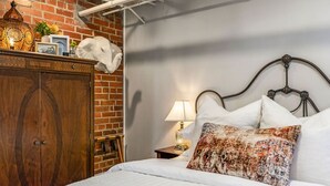A bedroom with a wooden wardrobe, a bed with white bedding, a patterned pillow, a nightstand with a lamp, and a brick wall.