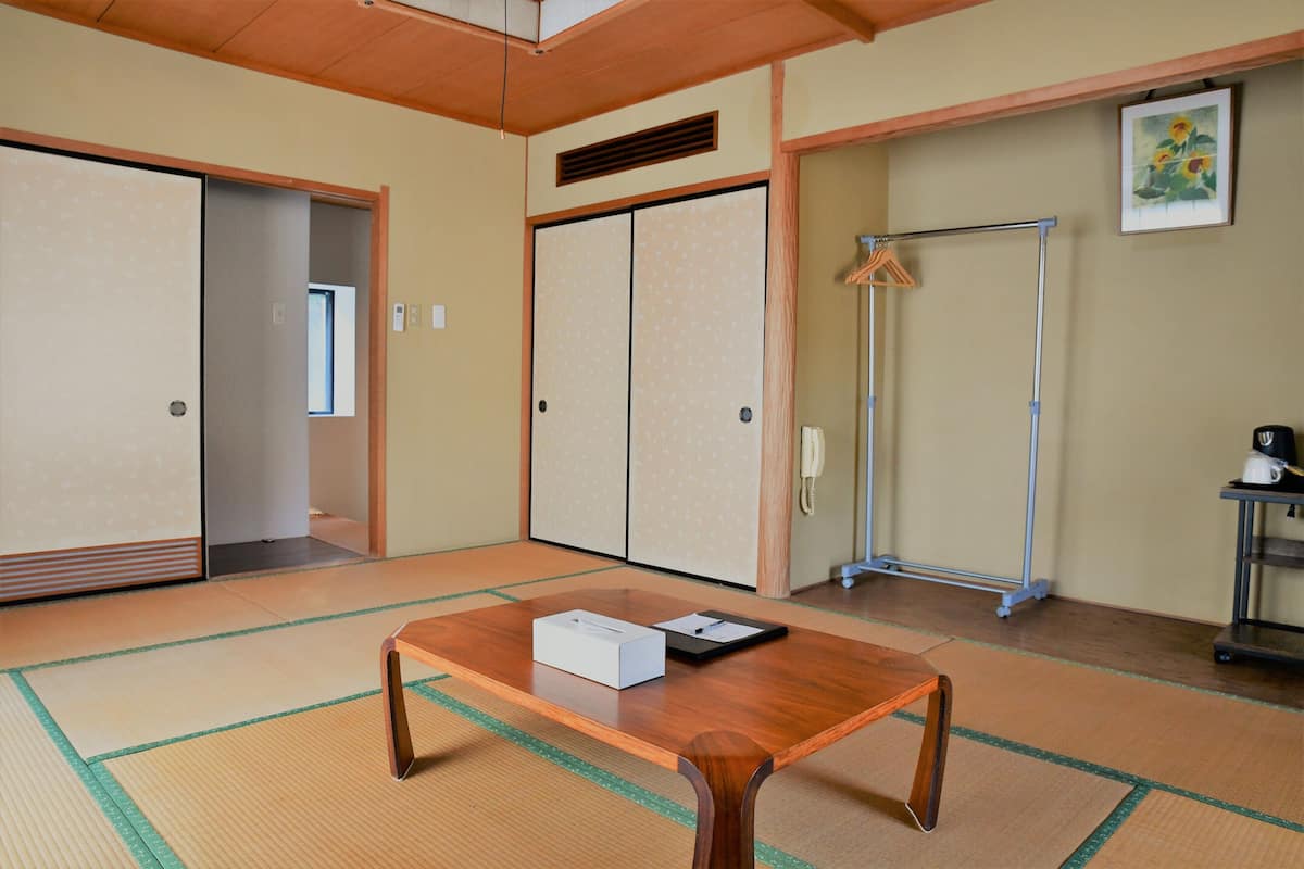 A traditional Japanese room with tatami flooring, a wooden table with a box and a black item, sliding doors, a hanging clothes rack, and a framed picture on the wall.