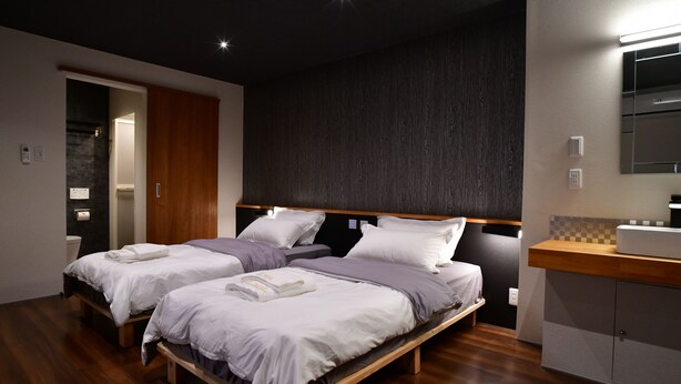 A modern bedroom with two beds, a wooden floor, a dark wall, and a small kitchenette. - Villa 33 Kashikojima (Shima)