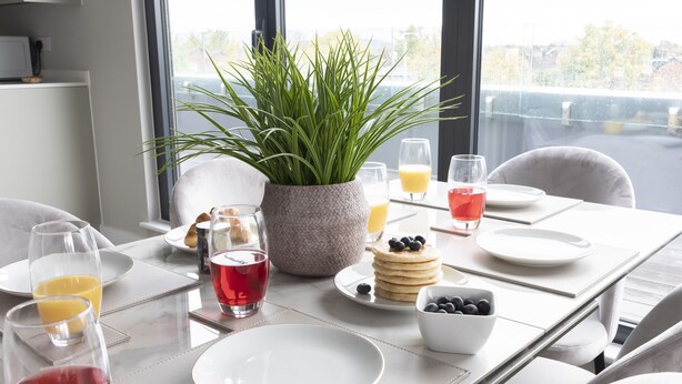 Een ontbijttafel gedekt met borden, glazen, een kommetje bosbessen, een plant en een stapel pannenkoeken.
