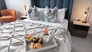 Een net opgemaakt bed met een dienblad ontbijtartikelen, een nachtkastje met een lamp en een stoel in een hotelkamer.