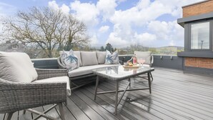 Een terras met rieten meubels en een glazen tafel, met uitzicht op een stadslandschap en heuvels.