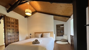 Une chambre spacieuse avec un grand lit, des poutres apparentes en bois au plafond, une bibliothèque intégrée et un coin lecture confortable.