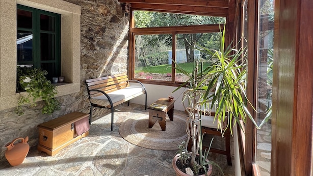 Une terrasse accueillante, avec un sol en pierre, des meubles en bois, un mur de pierre et une grande fenêtre donnant sur un paysage verdoyant.