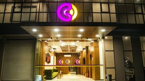 Entrada de hotel con un logotipo destacado, una recepción y una escalera que sube a un nivel superior.