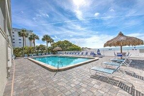 Ein Swimmingpool mit Liegestühlen und einem Strohdachschirm, umgeben von Palmen und einem Gebäude im Hintergrund.
