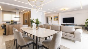 A modern living room with a dining area, a sofa, a TV, and a kitchen in the background.