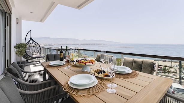 A dining area with a wooden table set for a meal, overlooking a scenic view of the sea and mountains.