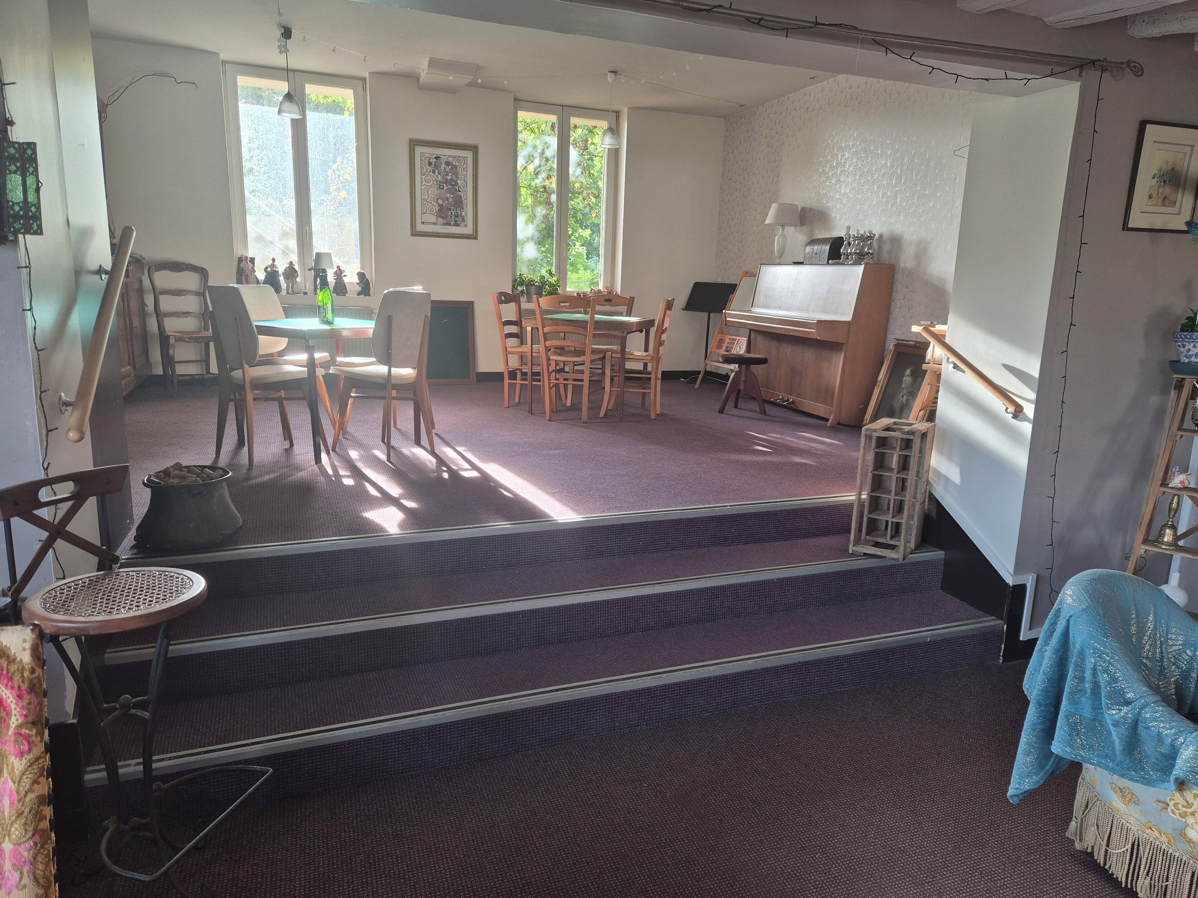 A room with a piano, a dining table with chairs, a staircase, and a window with a view of trees.